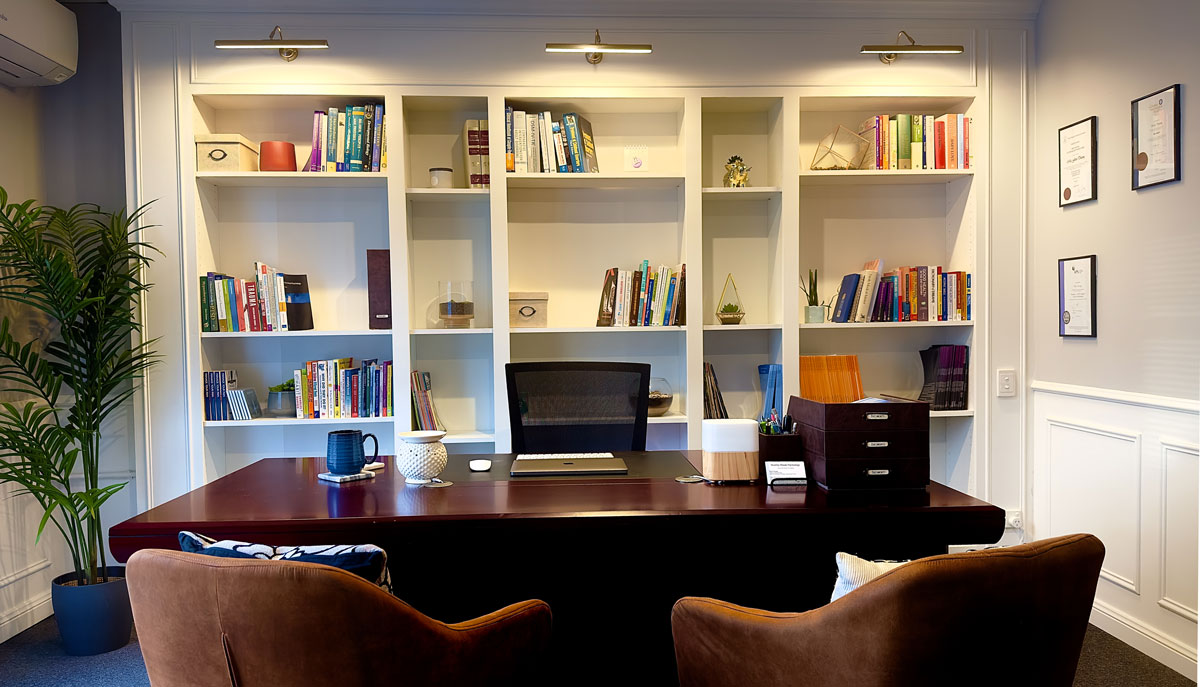 Interior photo of consultation room, tidy wooden desk, chairs and bookshelf in the background